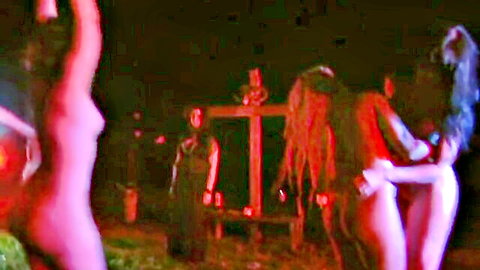 Nude dreadlocked women with exposed tits surround man in dark red-lit horror ritual