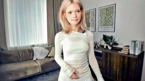 Blonde webcam girl in sheer white long-sleeve dress teasing with smile in cozy living room