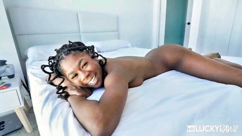 Nude smiling ebony babe with braids lying sideways on bed showing perky tits and ass