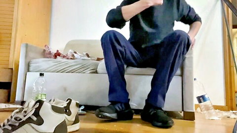 Gay man sits spread-legged on couch showing off black sneakers for foot worship fetish