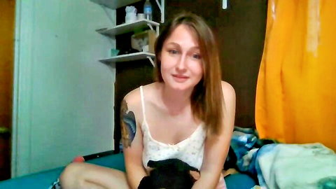 Brunette webcam girl in camisole smiling while holding black cat on bed