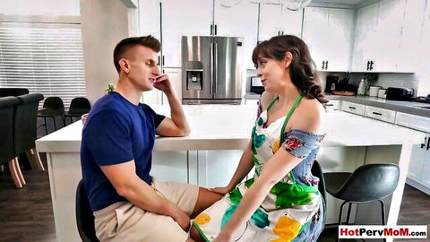 Curvy brunette stepmom in floral apron dress flirts smiling with stepson at kitchen island