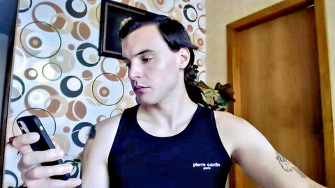 Handsome guy with ponytail in penis tank top holds phone selfie in bedroom