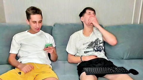 Two gay twinks in shorts laugh on couch during webcam chat