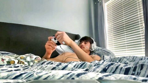 Shirtless twink gamer boy lying on bed playing Xbox controller with headphones smirking seductively