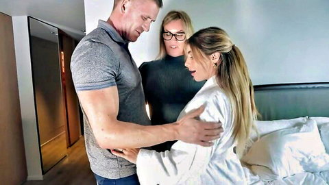 Muscular hunk embraces blonde babe in white robe as mature blonde watches smiling in hotel bedroom