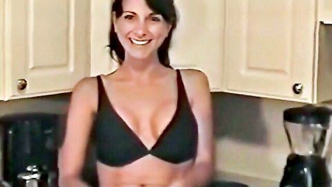 Brunette stepmom teases with deep cleavage in black top, smiling in kitchen