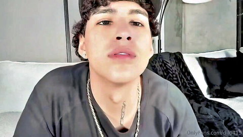 Handsome young curly-haired Latino guy smirking in bed wearing gray tee and gold chain