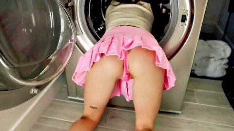Young beauty in pink pleated skirt bent over washer, exposing pink panties