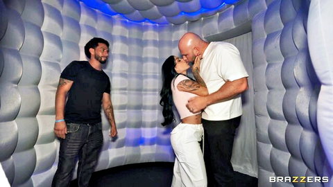 Muscular bald man passionately kissing tattooed brunette woman in photo booth while another guy watches