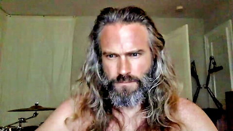 Shirtless muscular bearded hunk with long gray hair stares intensely at webcam