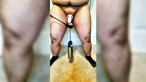 Chubby bear stands naked with pink strap-on dildo strapped over his exposed cock and balls above metal flask on floor