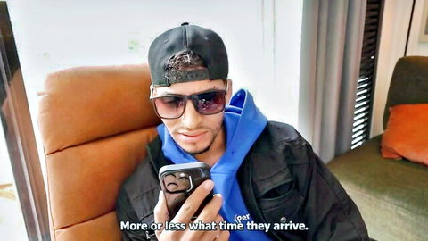 Tanned man in cap sunglasses and hoodie checking phone on couch waiting for arrival