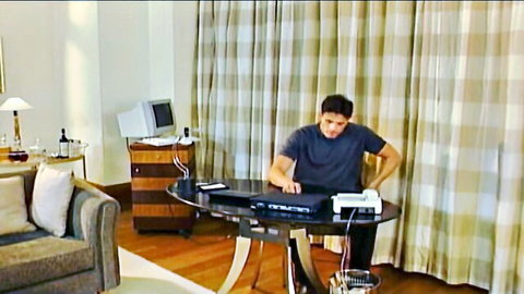 Handsome dark-haired man in t-shirt typing on laptop at desk in luxury hotel room