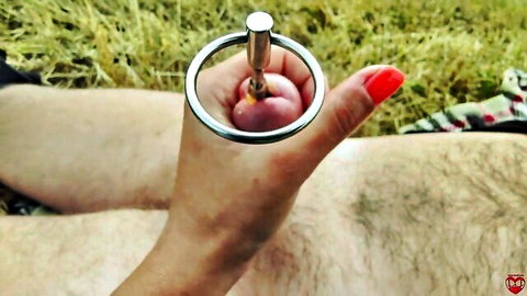 Red-nailed hand inserts steel urethral sound into swollen cock glans outdoors in grass