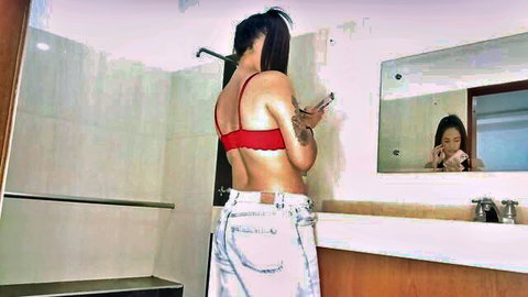 Tattooed brunette in red bra and low-rise ripped jeans poses for mirror selfie in bathroom
