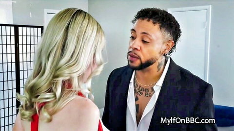 Blonde in red dress stares intensely at suited black stud with beard and tats, interracial tease.