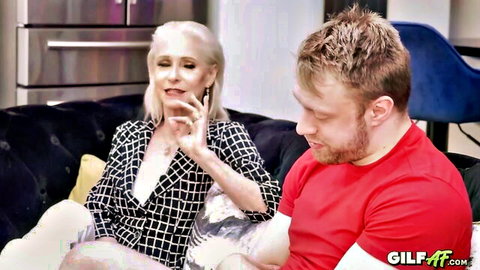 Blonde GILF in checkered blouse teases cleavage chatting intimately with young bearded hunk on couch