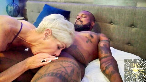 Mature blonde MILF in purple lingerie kisses tattooed black man's inner thigh on bed
