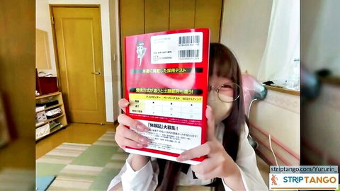 Cute Japanese amateur in glasses holds red manga book over face teasing in room