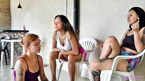 Three tattooed busty women in tank tops and shorts lounging dominantly on chairs in femdom tease