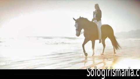 Blonde woman riding brown horse on sunny beach in white outfit