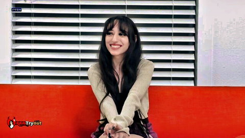 Young smiling brunette in open beige top sitting on red couch teasing viewers