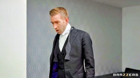 Blonde man in dark suit and white shirt stands looking intense in modern room