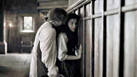 Man passionately embraces clothed woman from behind in dimly lit historical bedroom scene