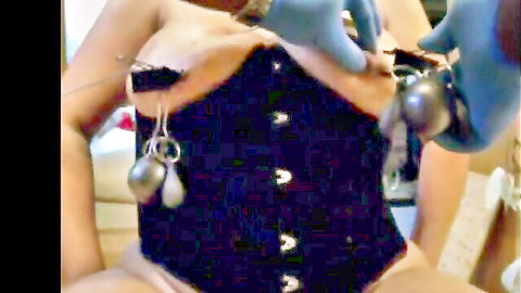 Woman in black corset screams as gloved hands insert needles into her large breasts with weights