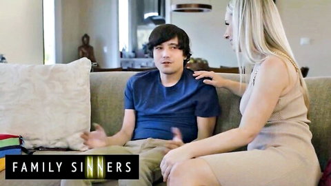Blonde MILF in beige dress touches young guy's shoulder seductively on couch