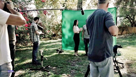 Curvy brunette MILF standing on outdoor green screen set surrounded by male film crew shooting porn video