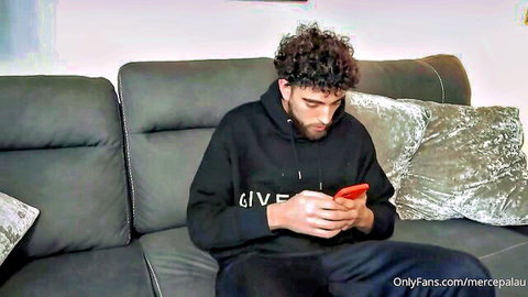 Handsome curly-haired bearded guy in black Givenchy hoodie sits on couch holding red phone teasing OnlyFans fans