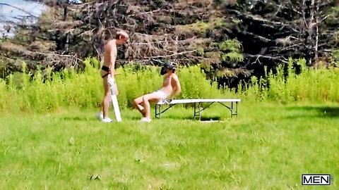 Two muscular gay hunks in tight bulging speedos outdoors, one spread-legged on picnic table