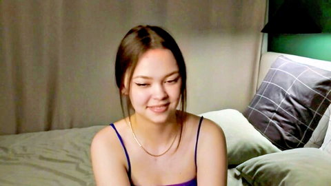 Cute Asian webcam girl smiling seductively in tight purple camisole on bed