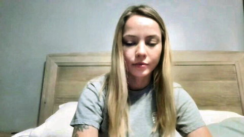 Blonde amateur webcam girl in gray t-shirt sitting on bed looking seductive