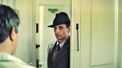 Handsome man in black fedora and suit stands in open doorway gazing intensely