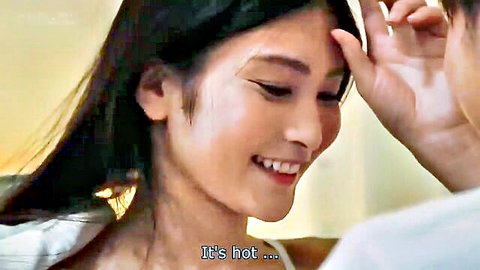 Seductive Asian brunette smiling saying it's hot touching her face in white top