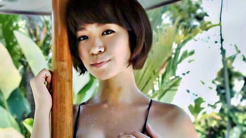 Busty Asian teen in black slip smiles leaning on pole in tropical vibe