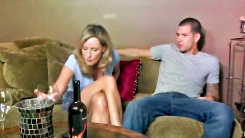 Lonely blonde cougar seductively pours wine for young stud on couch