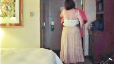Brunette MILF in white tank top and beige skirt holding red dress in hotel room closet