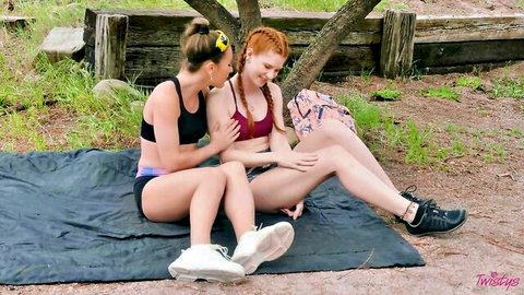 Redhead and brunette lesbians seductively touching thighs outdoors on blanket in bras and shorts