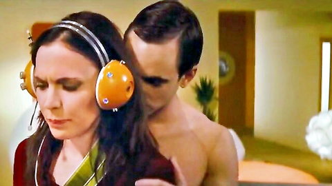 Lucy Egerton topless with headphones, man embracing from behind in nude scene