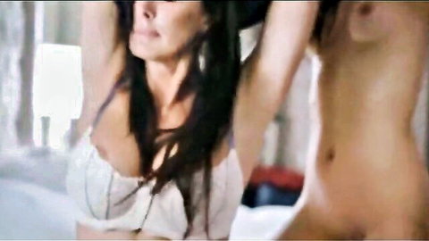 Two busty brunettes, one in sheer lace bra and one topless, raise arms exposing tits in seductive bedroom striptease