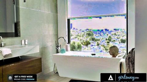 Blonde teen relaxing naked in bubbly bathtub with city view from luxurious bathroom