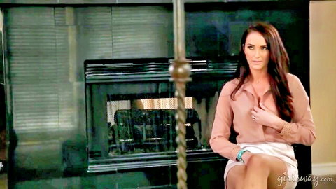 Sexy brunette MILF in pink blouse and white skirt sits teasing with legs crossed by fireplace