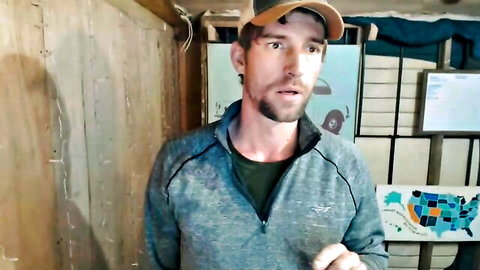 Bearded man in gray zip-up jacket and cap speaking in dimly lit wooden room with US map poster.