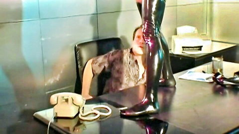 Woman in shiny latex boots dominates man at desk in fetish office tease