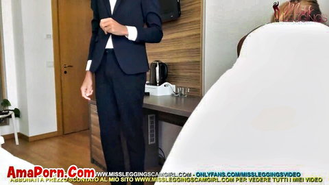 Suited businessman stands in hotel room eyeing sexy woman on white bed sheets