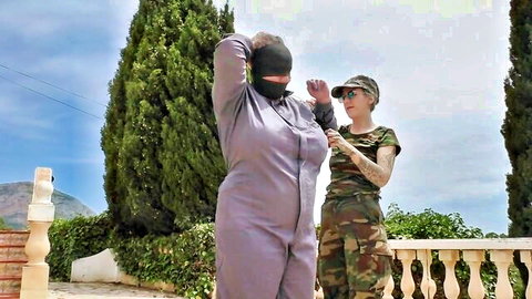 BDSM slave in hood submissive pose with camo dominatrix outdoors